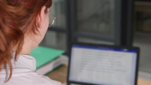 Woman in Glasses Looks Into Laptop Screen While Working at Computer Sitting at Desk in Office