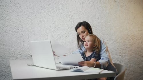 Young Mother with Headphone Working on Laptop. Her Crying Child Sits Woman's Lap