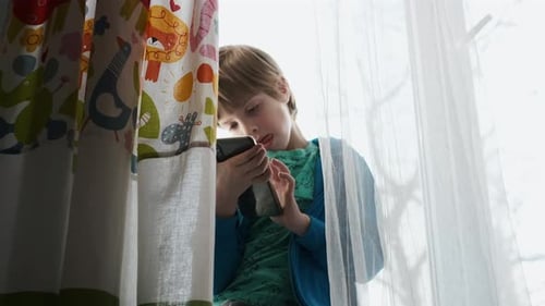 Boy Sits at Window Sill Looking at Phone
