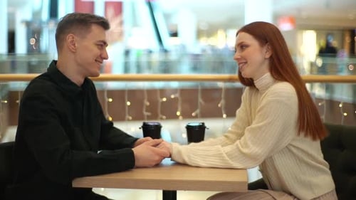 Side View of Beautiful Young Couple Drinking Coffee Holding Hands and Talking Looking at Each Other