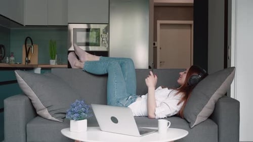 Faule kaukasische Frau auf Kopfhörern, die auf dem Sofa liegt und Smartphone oder Handy in der Moderne benutzt