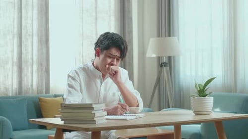 Asian Man Student Being Sleepy While Writing In The Notebook On The Table At Home