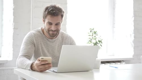 Man Using Phone and Laptop in Bright Office