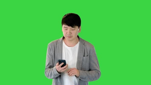 Young Asian Man Walks Looking at His Smartphone on a Green Screen Chroma Key