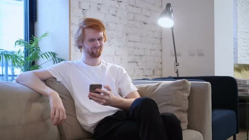 Red-Haired Man Reacts to Phone on Couch
