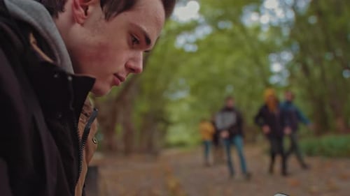 Young Guy with a Phone in His Hands Sitting on a Bench in the Park. In the