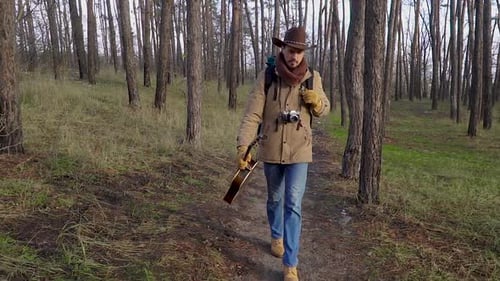 A Man with a Guitar and a Camera Walks Along a Trail in the Forest
