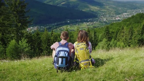 Young Couple of Tourists with Backpacks Sitting on a Green Meadow, Looking at a Picturesque Mountain