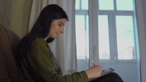 Woman Writing in Notebook at Home by Window