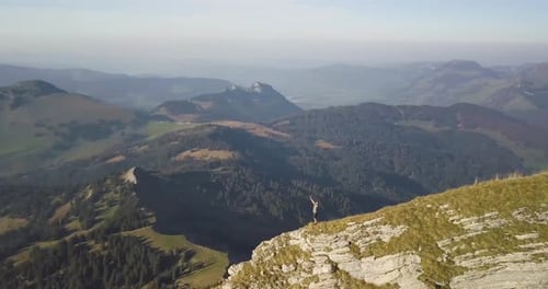 A hiker stands on the edge of a mountain ridge and puts is hands in the air.