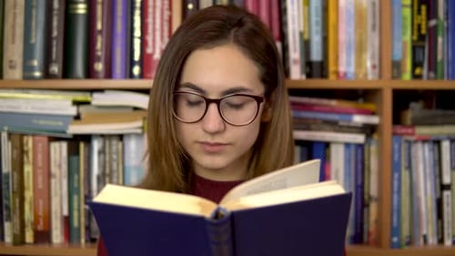 A Young Woman Is Reading a Book in a Library. A Woman with Glasses Carefully Looks at the Book