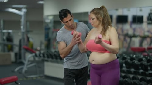 Fit Male Personal Trainer Supporting Overweight Sportswoman Training with Dumbbells in Gym