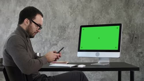 Businessman Looking at His Smartphone Messaging and Sitting Near Computer Screen. Green Screen Mock