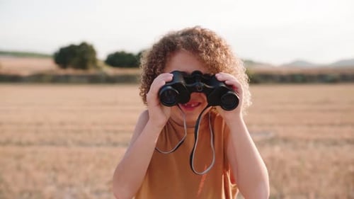 Cute boy looking through binoculars and watching wilderness and animals on agricultural field,