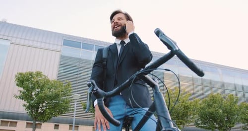 Gerente de oficina masculino con barba sentado en su propia bicicleta cerca de un gran edificio moderno