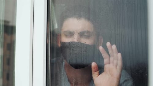 Sad man in medical mask walks window wet after rain look outside during quarantine due coronavirus
