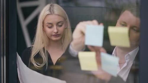 Jeune femme concentrée parlant avec un homme écrivant sur des notes adhésives sur une porte vitrée