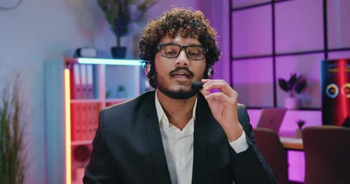 Indian Businessman in Headset Talking Into Camera on Evening Office Room Background