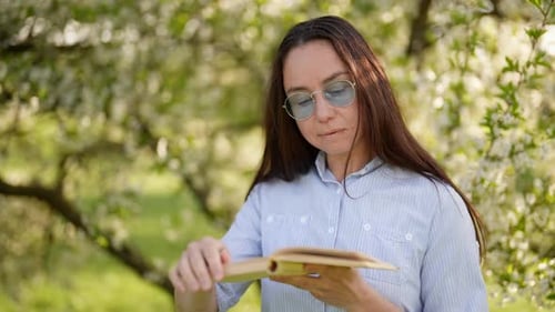 Calm Woman is Reading Book in Blooming Garden in Sunny Spring Morning Portrait of Lady with Glasses