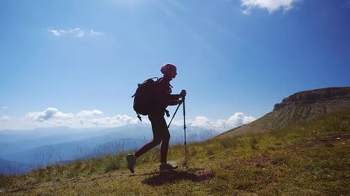 Young Woman with Backpack and Trekking Poles Walks Uphill Towards the Summit Against Blue Sky. Slow