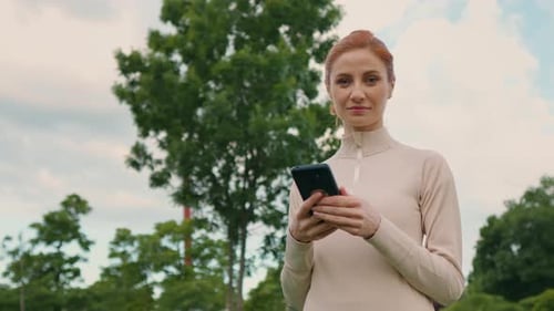Woman portrait in park using smartphone looking to camera while morning jogging.