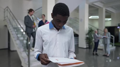 Young Intelligent African American Man Analyzing Paperwork Standing in Office Lobby with Colleagues