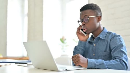 Young African Man with Laptop Talking on Smartphone in Office