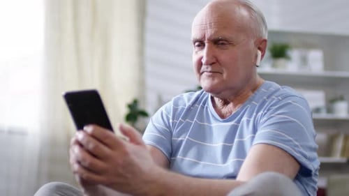 Senior Man Using Smartphone with Earbuds Indoors