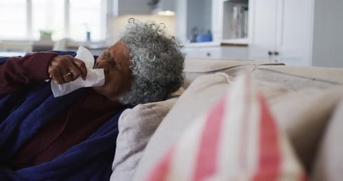 Une femme afro-américaine âgée malade couverte d'une couverture éternuant alors qu'elle était allongée sur le canapé à la maison
