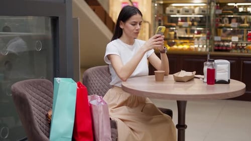 Woman Sending Photo of Her Purchases From Smartphone to Friend While Sitting in Cafe After Shopping