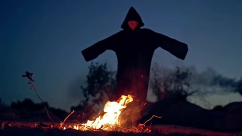 Scary witch stands by the fire at night. Black figure in cloak with hood walking around the fire wit