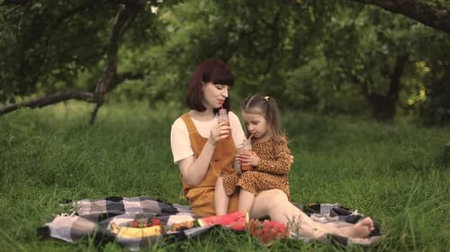 Young Mom with Her Little Cute Daughter on a Picnic in Green Park Drinking Fresh Juice