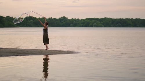A Young Female Artist Shows a Soap Bubble Show Blowing Up Huge Soap Bubbles on the Shore of a Lake