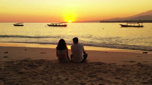 Young couple in love sitting on tranquil exotic beach, watching beautiful sunset with orange sky ref