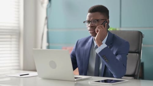Angry Young African Businessman Talking on Phone While Using Laptop in Office