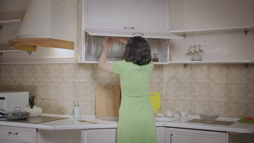 Girl in a Dress Puts Things in Order in the Kitchen Cabinet Arranges the Dishes Rear View