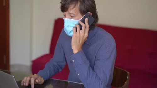 A Young Man Wearing a Face Mask Works From Home During Coronavirus Self-isolation. He Talks on a