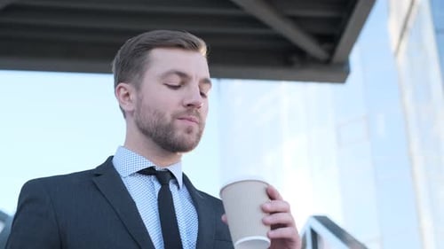 Young Thoughtful Man Standing Outside Modern Building and Drinking Coffee