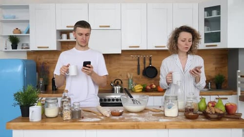 Couple Standing at Counter, Using Smartphones and Baking