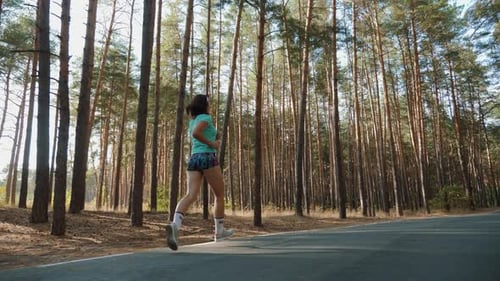 Sportive Woman Runs Along the Road in a Pine Forest