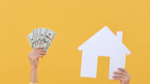 Holding Money and a House Cutout Against Yellow Backdrop