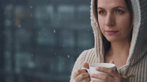 Caucasian Woman Stays on Balcony During Snowfall with Cup of Hot Coffee or Tea
