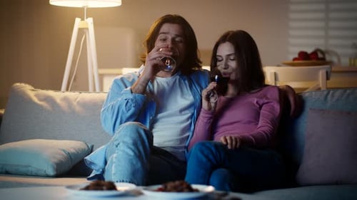 Young Contented Man and Woman Sitting on Sofa with Wine Glasses and Embracing Watching Tv Together