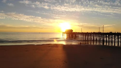 Drone flying under the Newport Beach pier at California sunset with sunlight beams and people in sil