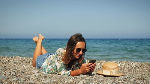 Cheerful charming brunette girl lying on beach and using apps on smartphone at windy summer day.
