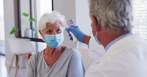 Male caucasian doctor measuring temperature of senior caucasian woman at hospital