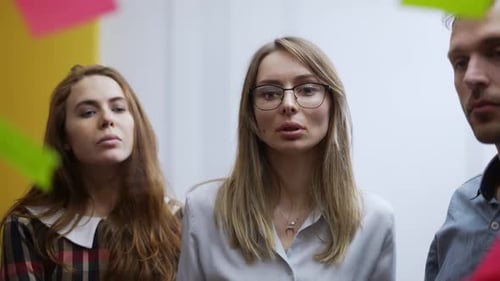Blonde Businesswoman on Team Meeting Sticks the Color Note on Glass Wall Front View