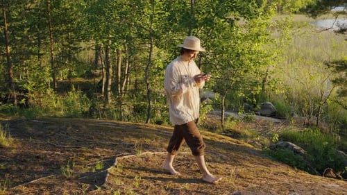Young Man Walks Barefoot on Bank and Looks at Phone Screen