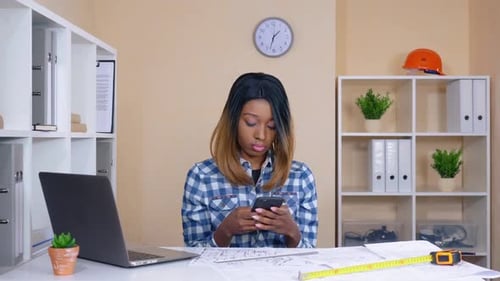 Woman Using Cell Phone at Desk with Plans