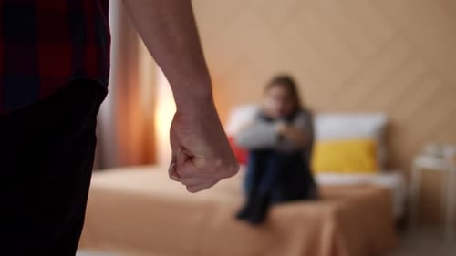 Husband with clenched fist goes to wife in fear sitting on bed, back view. Man clenches fist front